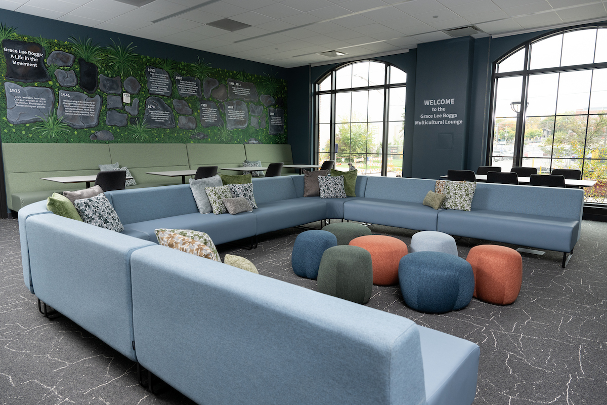 A modern lounge features light blue sectional sofas, colorful round ottomans, and a wall mural depicting the life of Grace Lee Boggs.
