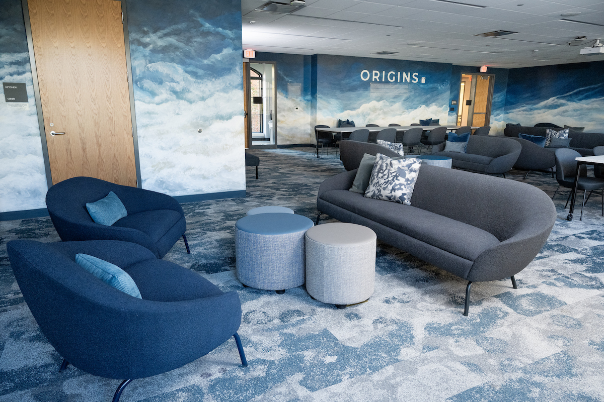 A modern lounge area with blue and gray seating, cloud-themed wall murals, and a "ORIGINS" sign.